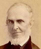 Portrait of an older man with a receding hairline, white beard, and formal dark jacket, looking directly at the camera.