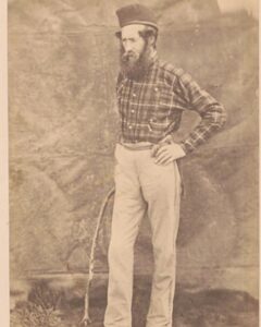 a photograph of a white man with his hand on his hip in a plaid shirt. He has a beard and wears a plaid shirt. the photo appears very old. 