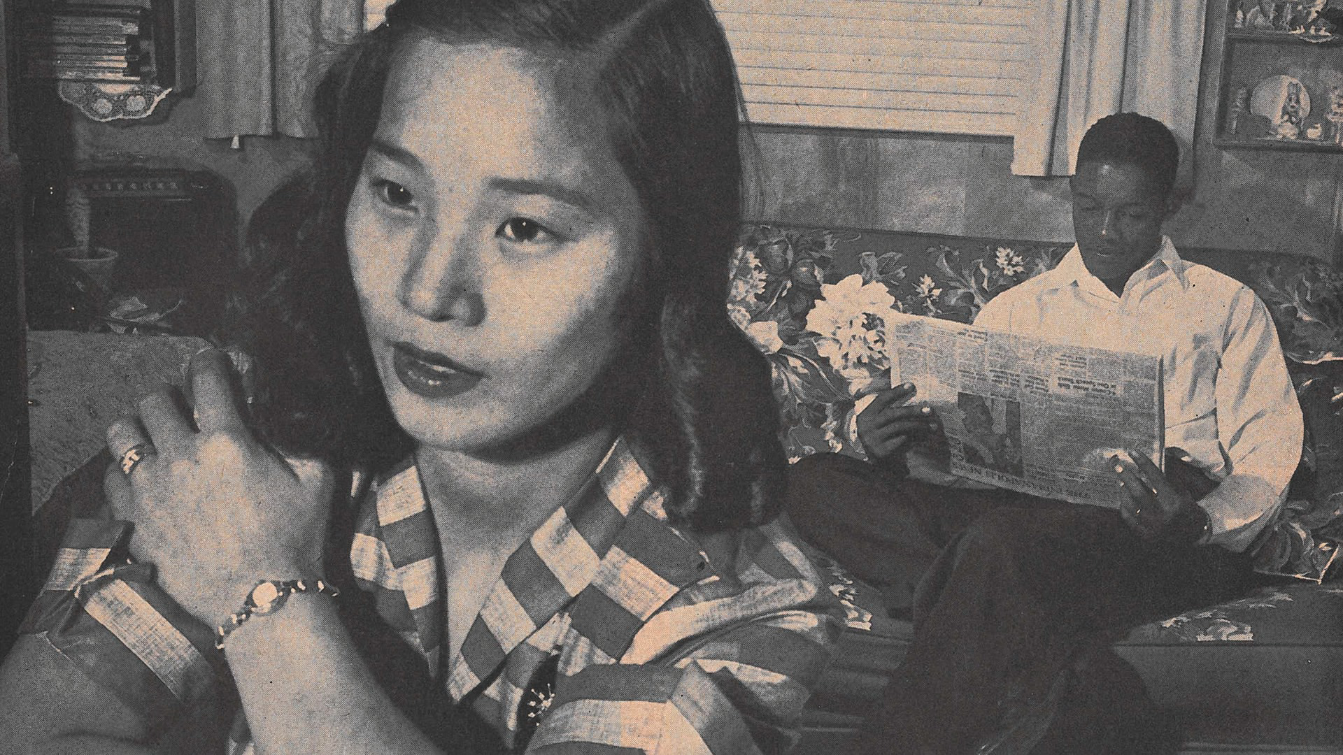 A Japanese woman stares out a window. A Black man sits behind her, reading the paper.